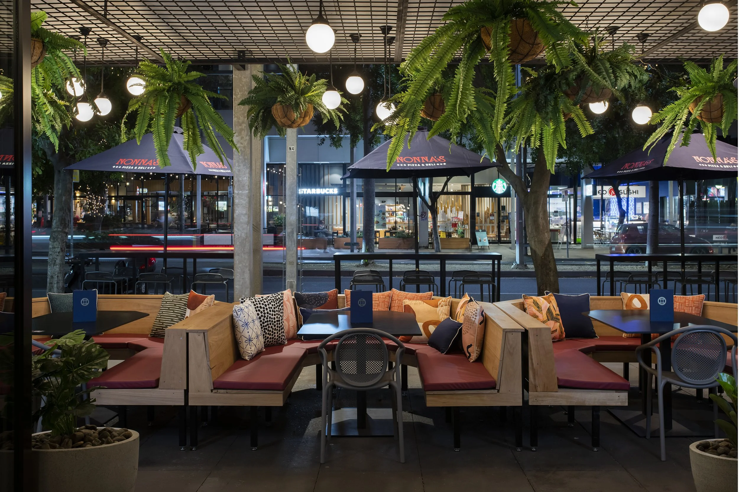 Outdoor restaurant seating area with wooden benches, colorful cushions, hanging plants, and ambient lighting, street view with storefronts and umbrellas in background.