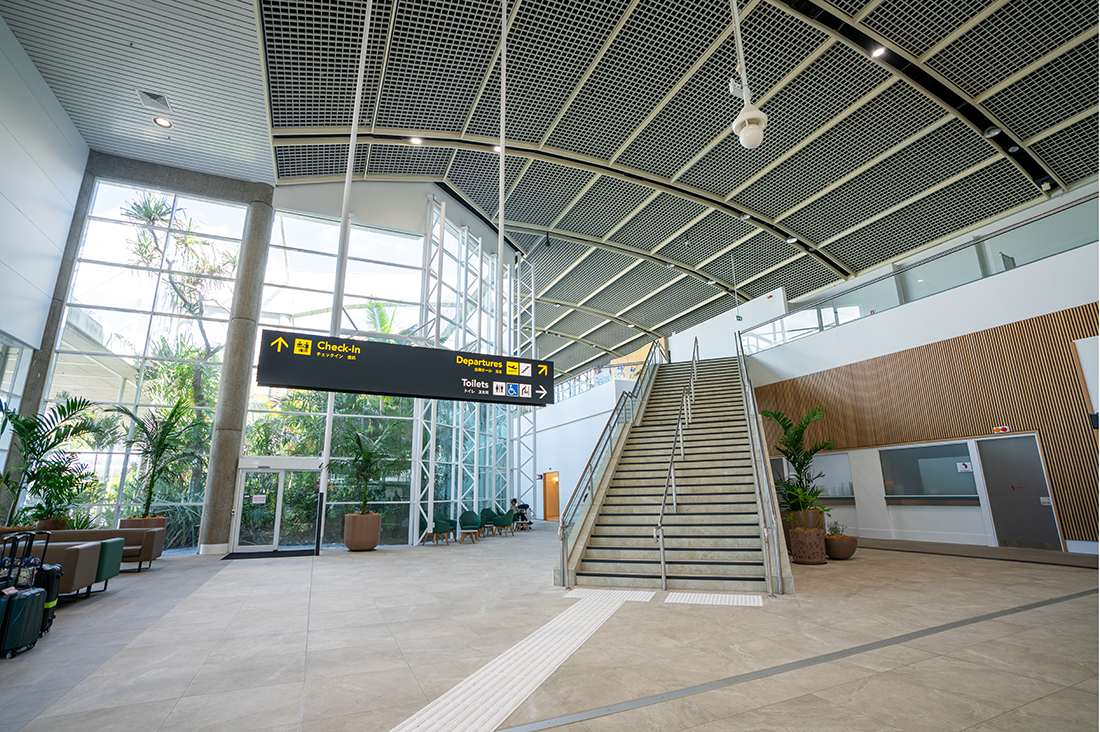 Cairns International T1 Airport. Modern airport terminal interior with a staircase, seating area, luggage, and directional signs for check-in and departures. Airport Interior Design Brisbane Australia