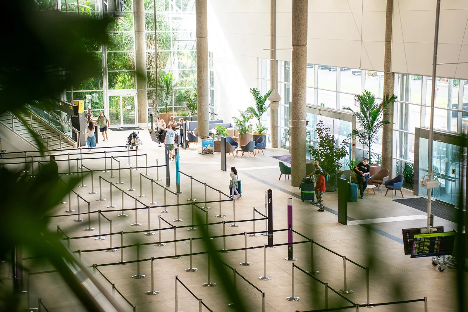 Interior of an airport terminal with seating area, greenery, large windows, and people waiting or walking with luggage.