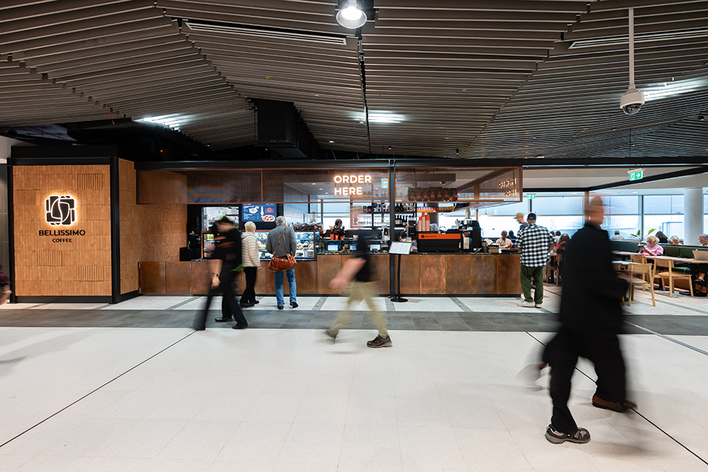 People walking and sitting in a modern coffee shop with "Bellissimo Coffee" sign and "Order Here" sign, ceiling with wooden panels, seating area with tables and chairs. Retail Aviation Interior Designers in Brisbane Australia