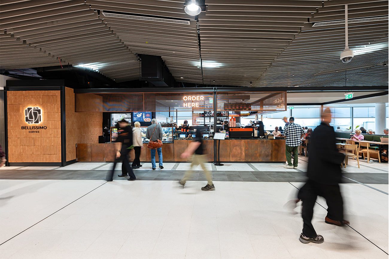 Interior of a coffee shop with a counter, people walking by, and a sign that reads 'Order Here'. Brisbane Airport Domestic Terminal. Retail Interior Design Australia