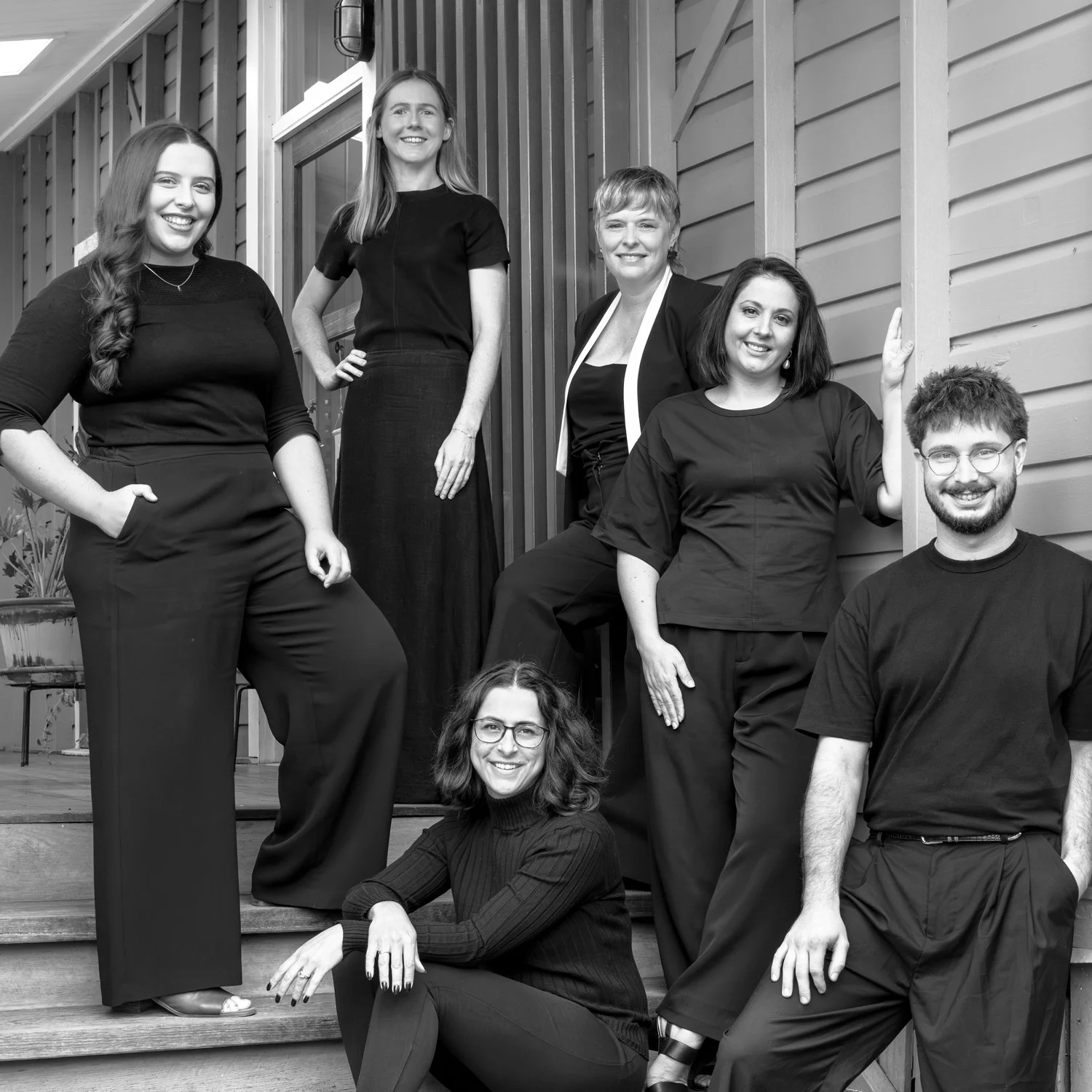 The studiospillane team,  posing on wooden steps outside a building. They are smiling and dressed in black or dark clothing.