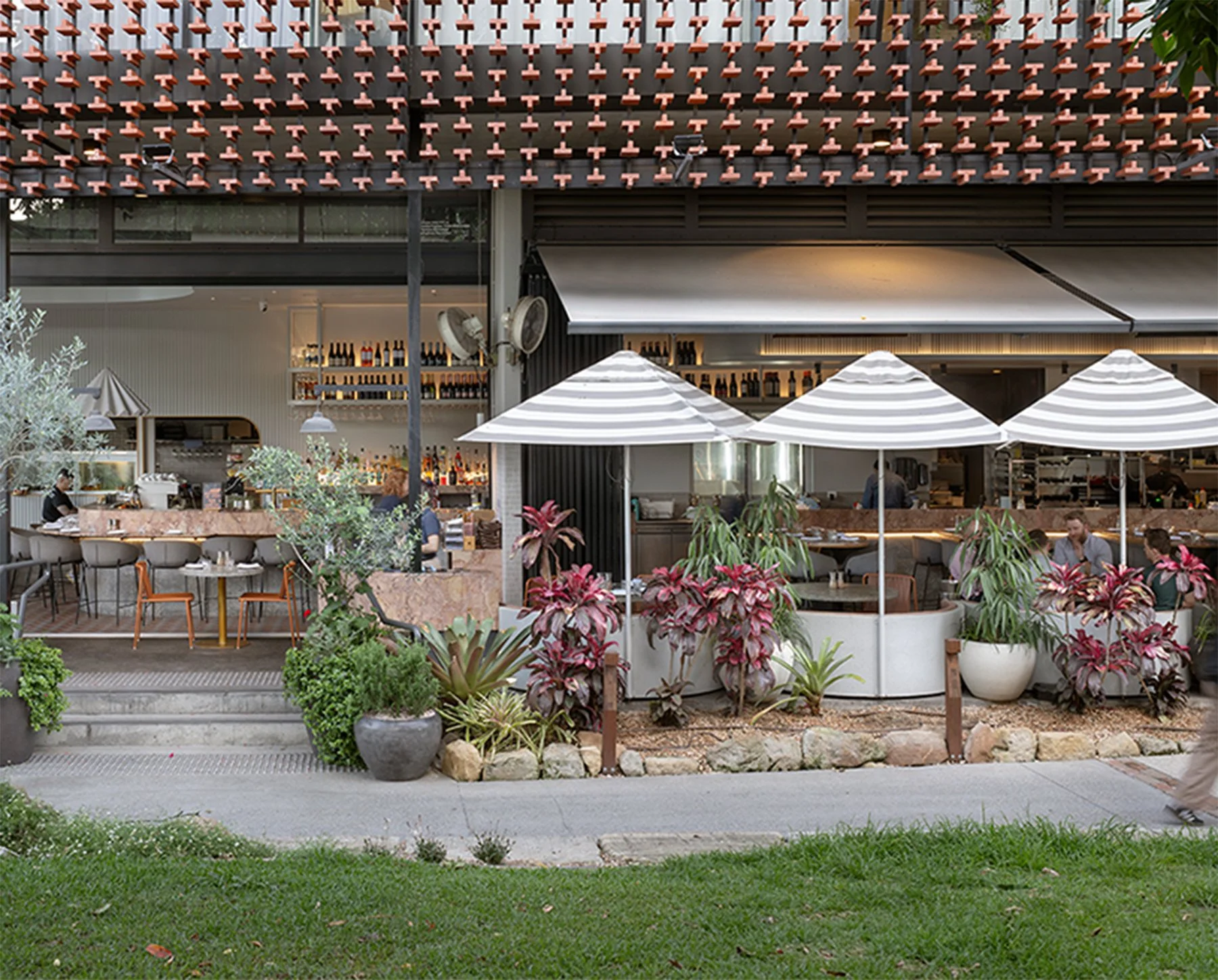 Outdoor restaurant patio with tables, chairs, and umbrellas, surrounded by potted plants and greenery.