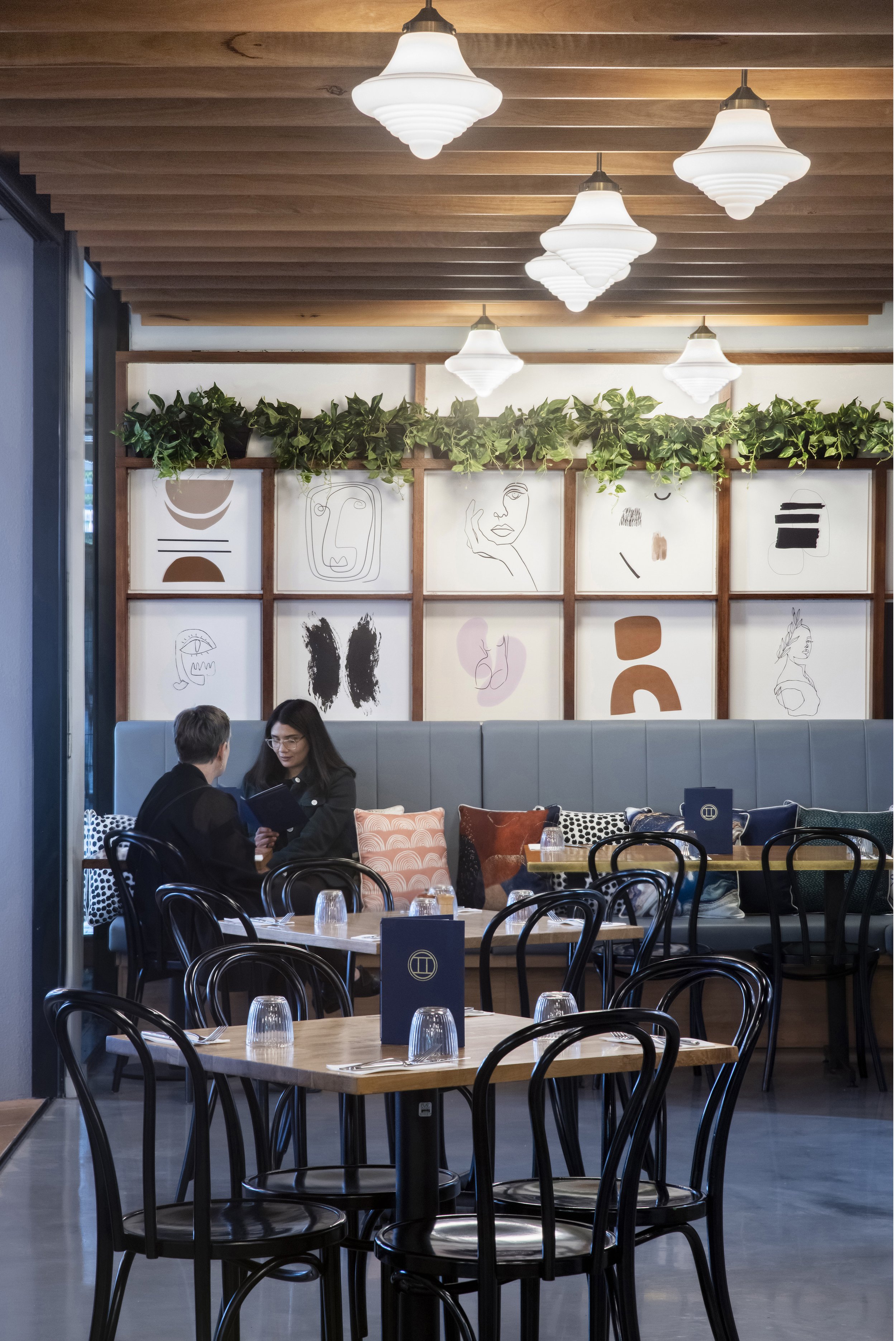Modern cafe interior with abstract art on the wall, plants, and pendant lights. Two people sitting at a table with menus and glassware, surrounded by cushioned bench seating and black chairs.