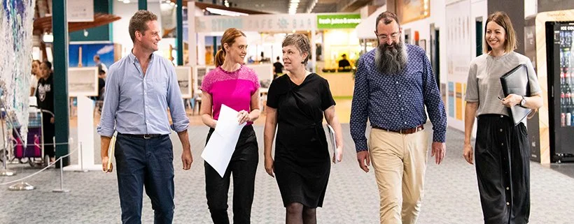 Five people walking and conversing in a modern indoor setting, likely at a convention or expo. They appear to be discussing ideas or planning, holding papers and folders. The background includes displays and a café area. Interior Designers in Brisban