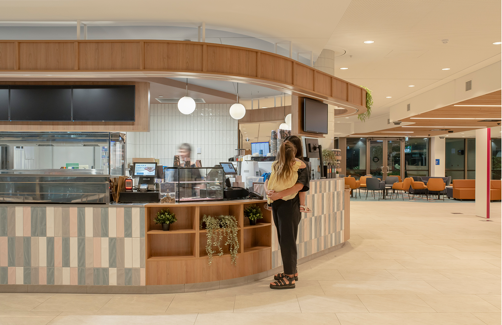 Prince Charles Hospital Foundation Common Good Cafe. A woman holding a child standing at a modern cafe counter with light wood decor, geometric tile design, and hanging round lights. Food and beverage Interior Design Brisbane Australia