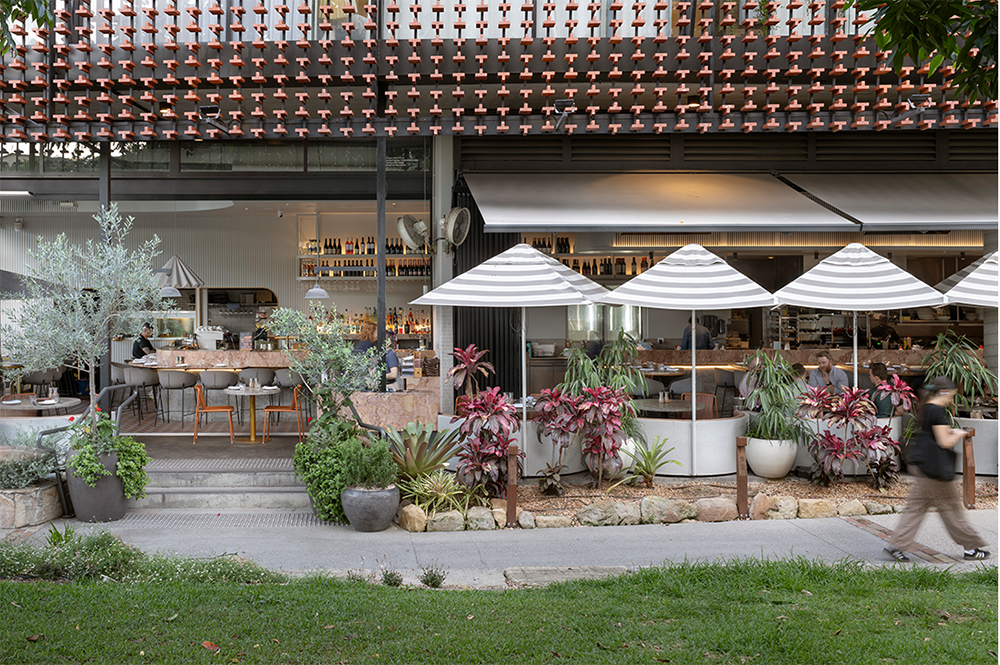 Outdoor café with striped umbrellas, greenery, and people seated at tables, viewed from across a grassy area.