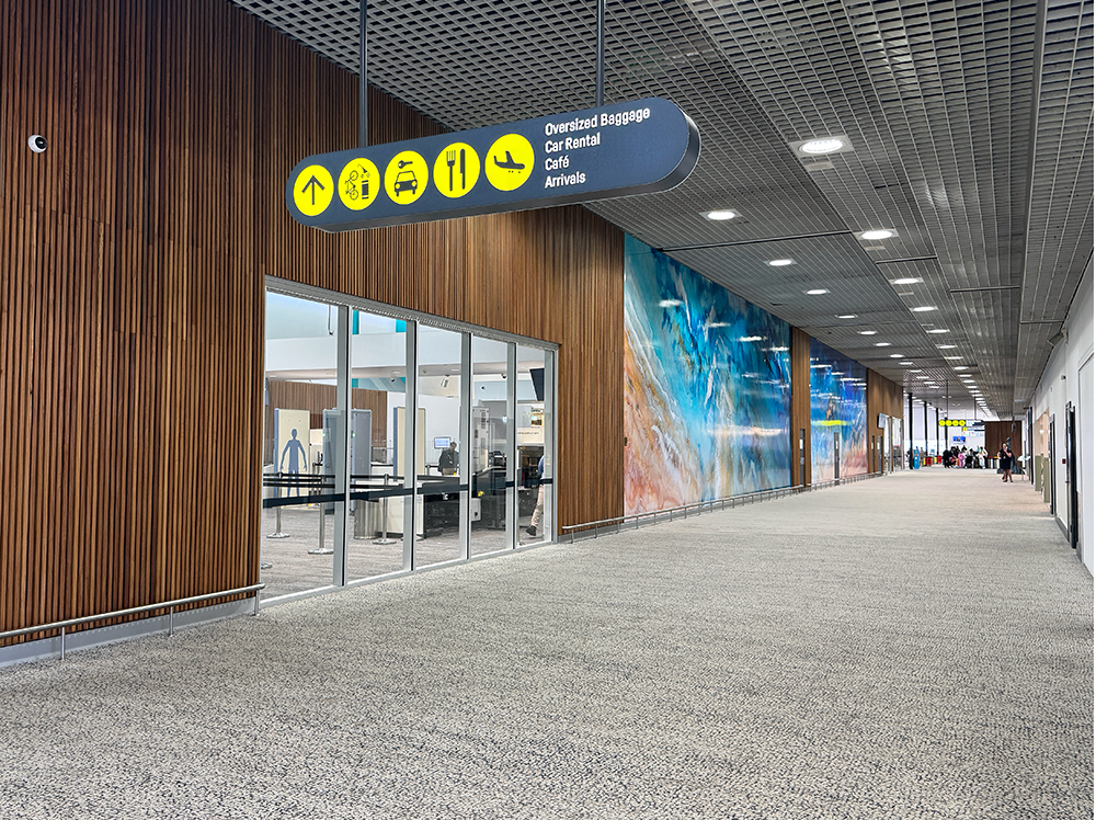 Airport corridor with signage for oversized baggage, car rental, café, and arrivals. Decor includes wooden wall panels and a colorful abstract mural. 
Townsville Airport. Interior Design Australia