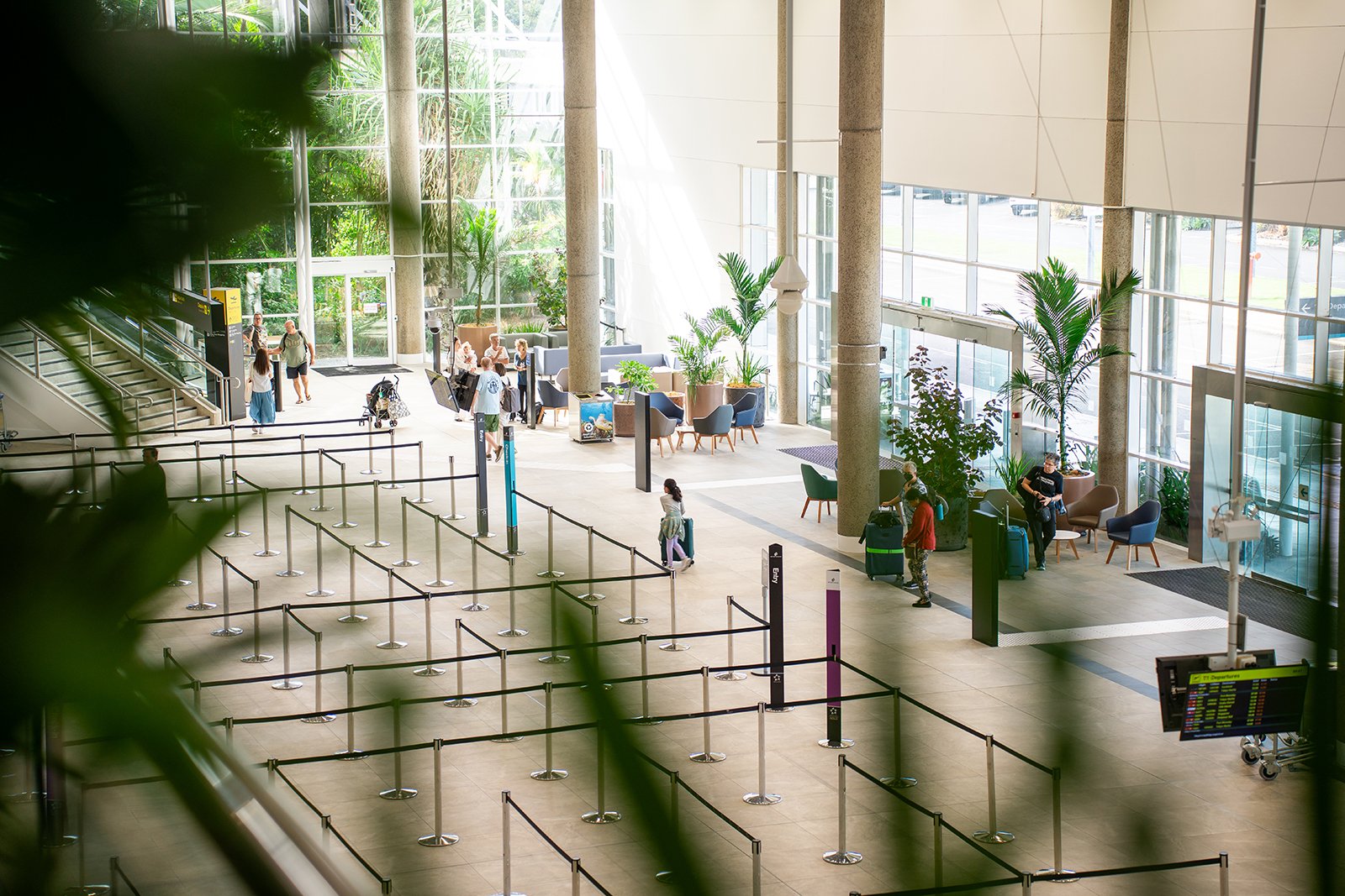 An airport waiting area with seating, people with luggage, large windows, and indoor plants.