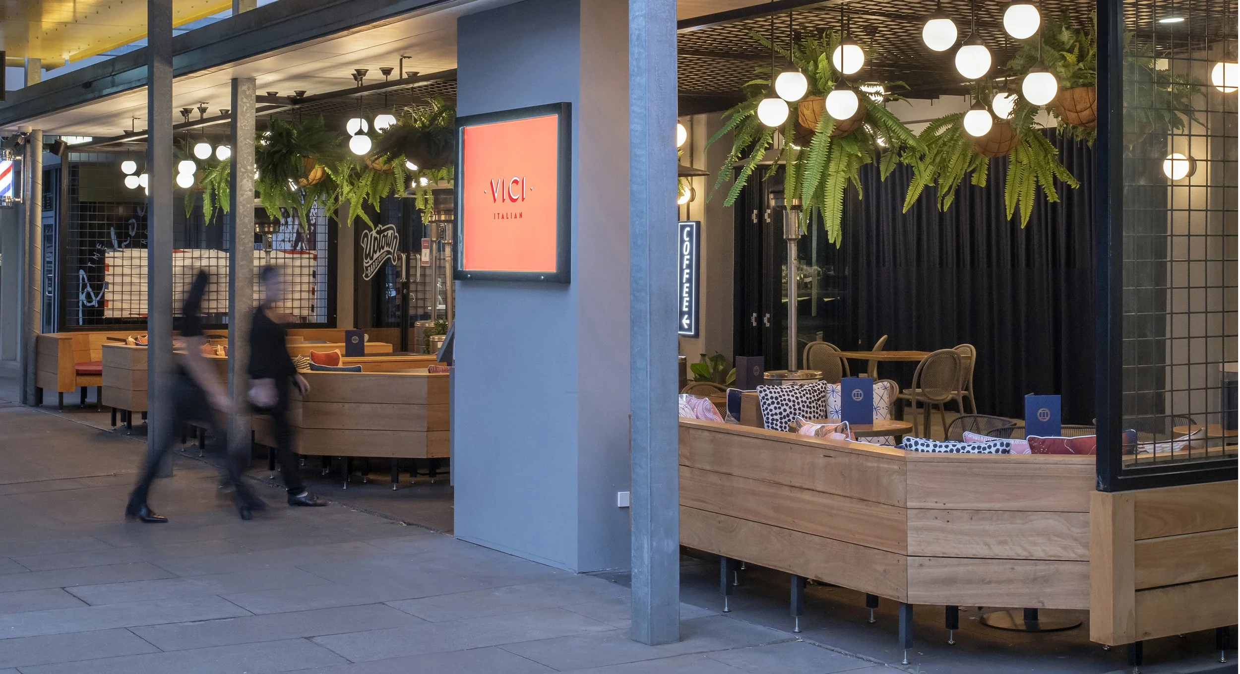 Outdoor seating area of a modern restaurant with wooden booths, decorative greenery, and hanging lights. People walk past the entrance where a sign reads "Vici Italian."