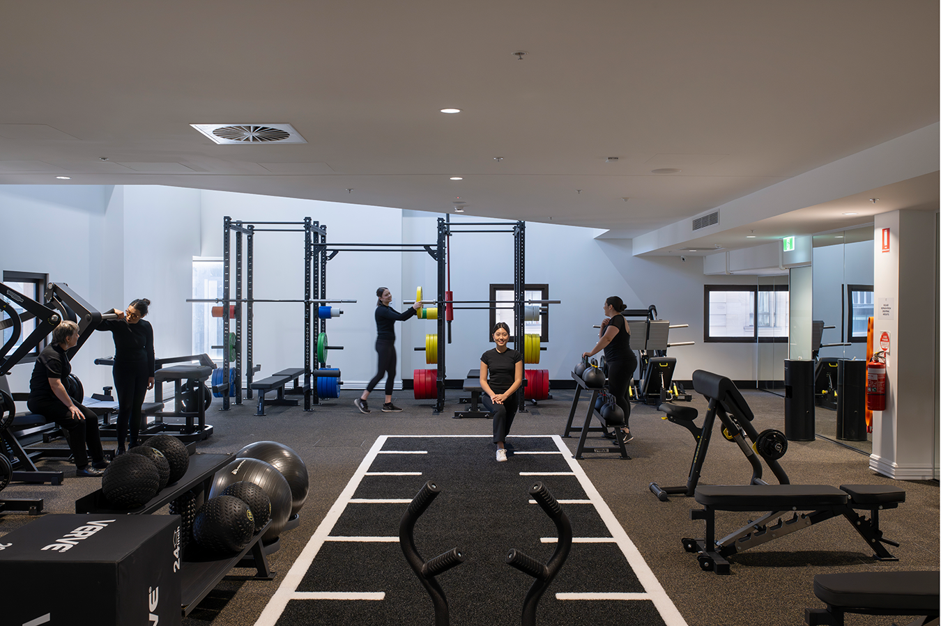 Tattersall's Club. Modern gym interior with people exercising using various fitness equipment, including weightlifting racks, benches, and exercise balls. Bright lighting and mirrored walls are present. Hospitality Interior Design Brisbane Australia
