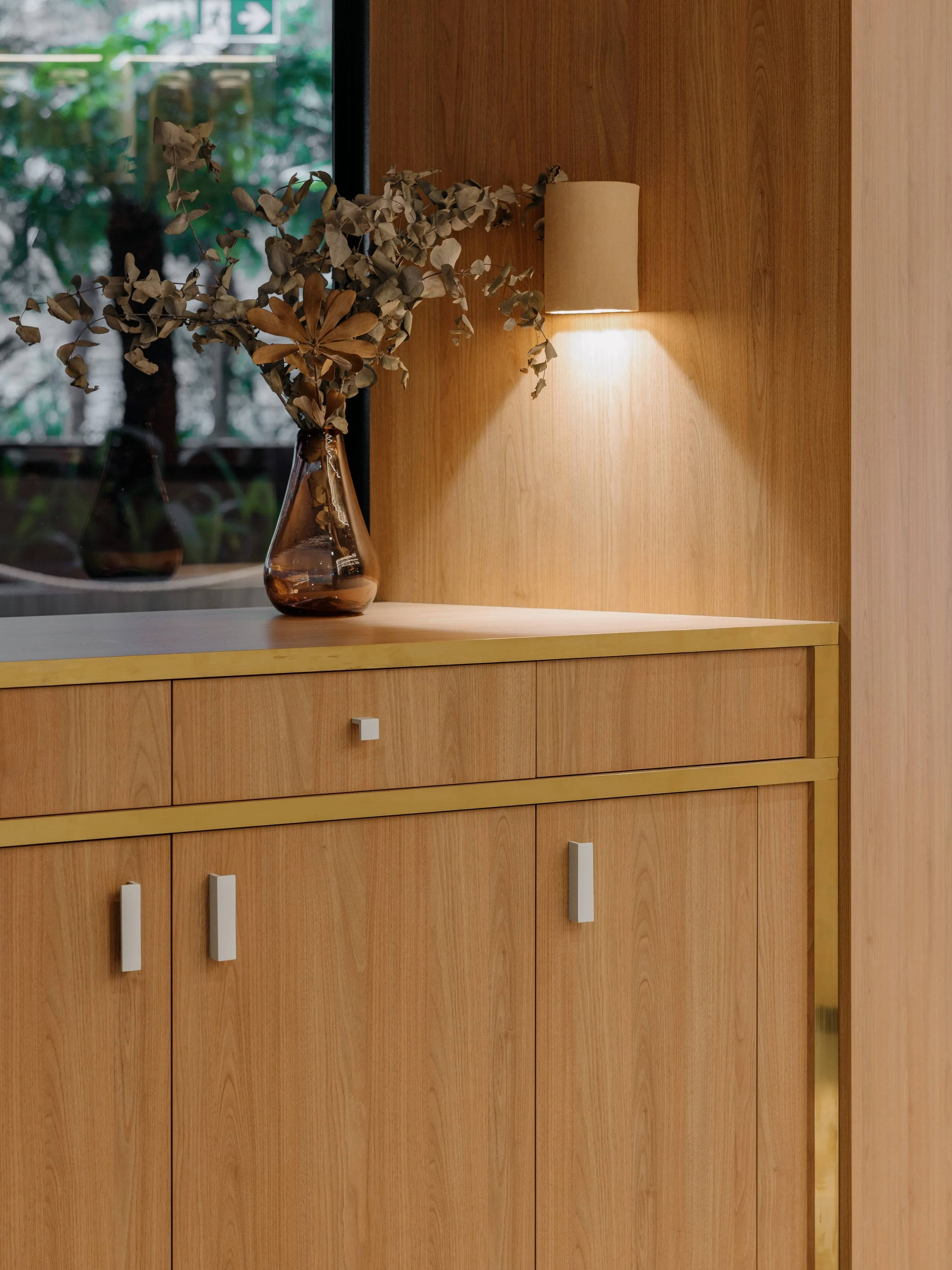 A wooden cabinet with white handles below a wooden wall. On top of the cabinet, there is a brown glass vase with dried leaves, and above it, a beige wall-mounted lamp casting light on the wall.