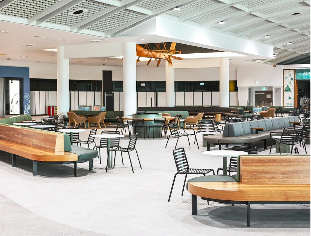 Empty interior space of an airport or shopping mall with modern seating, round tables, and a wooden boat hanging from the ceiling.