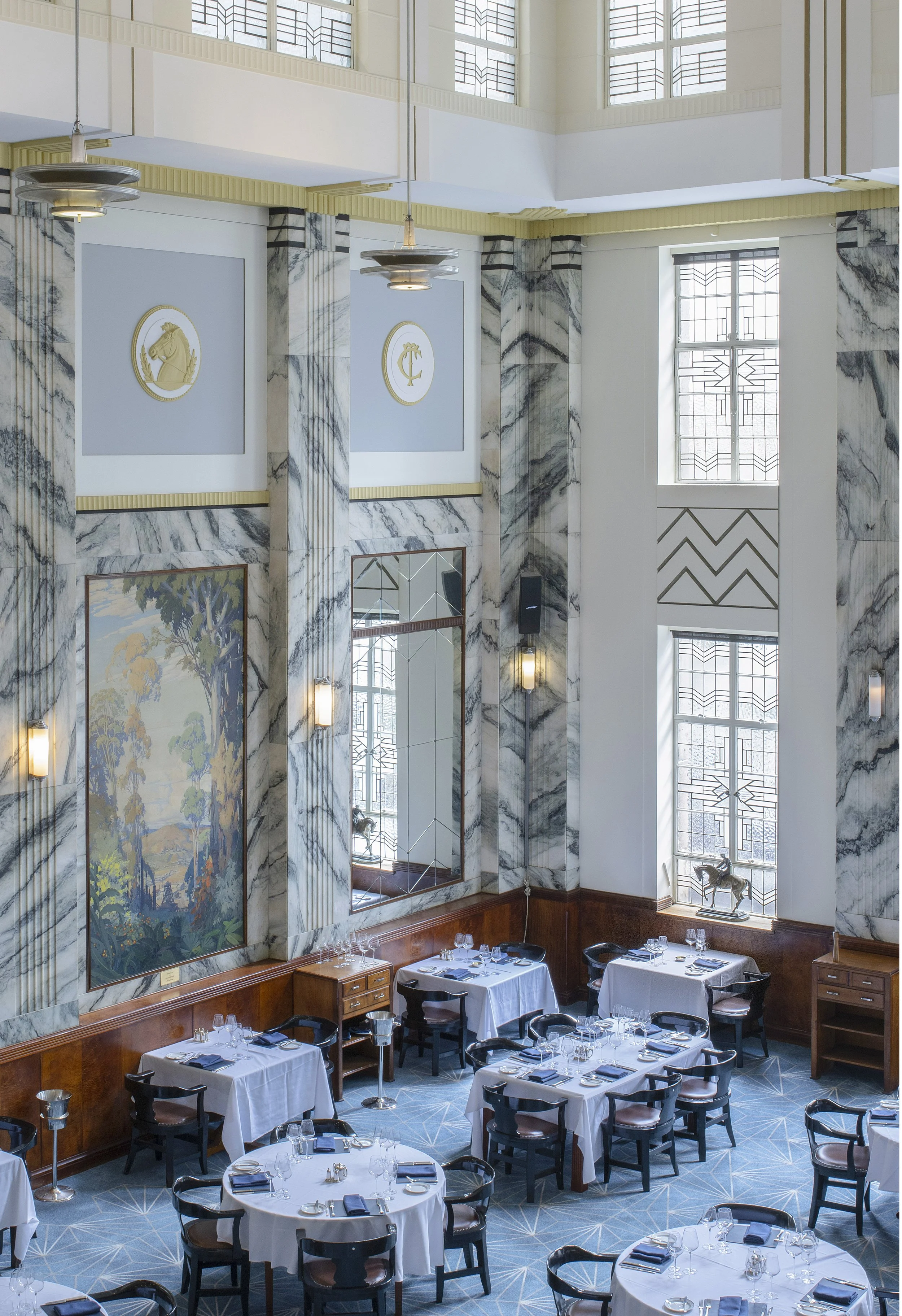 Art Deco styled dining room with round tables, white tablecloths, and elegant decor, including wall art and large windows.