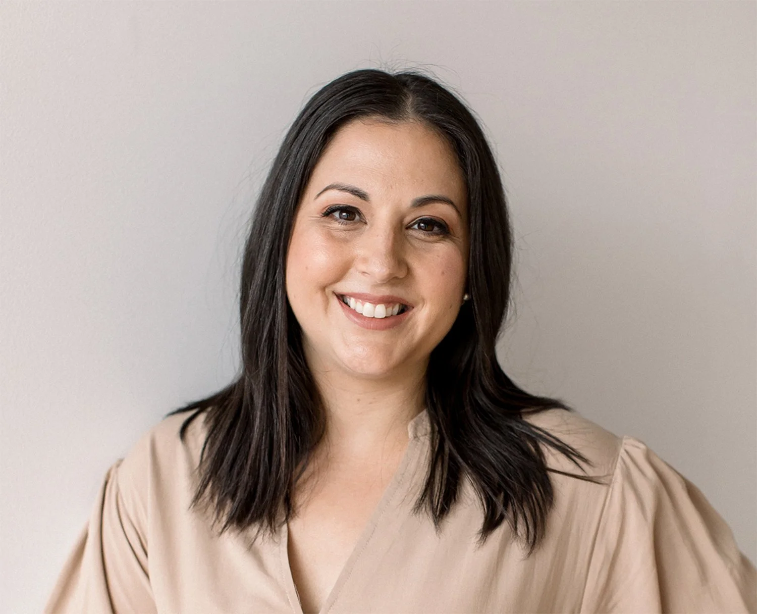 A woman with dark, shoulder-length hair smiling, wearing a beige top, standing against a plain wall.