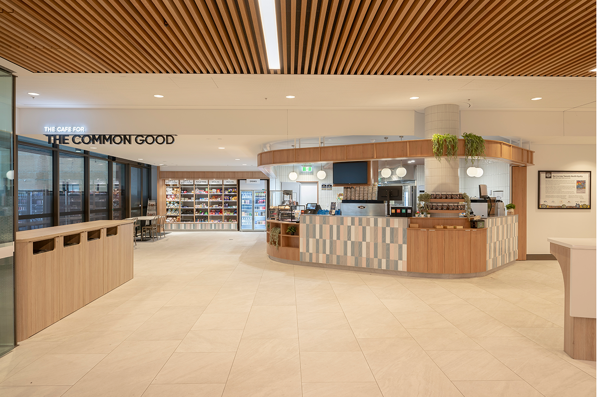 Prince Charles Hospital Foundation Common Good Cafe. Modern cafe interior with seating, snack shelves, and a tiled counter. Sign reads "The Cafe for the Common Good." Plants provide decor. Food and beverage Interior Design Brisbane Australia