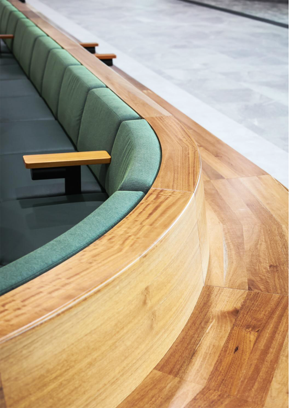 Curved wooden seating with green cushions and wooden armrests in a modern interior space.