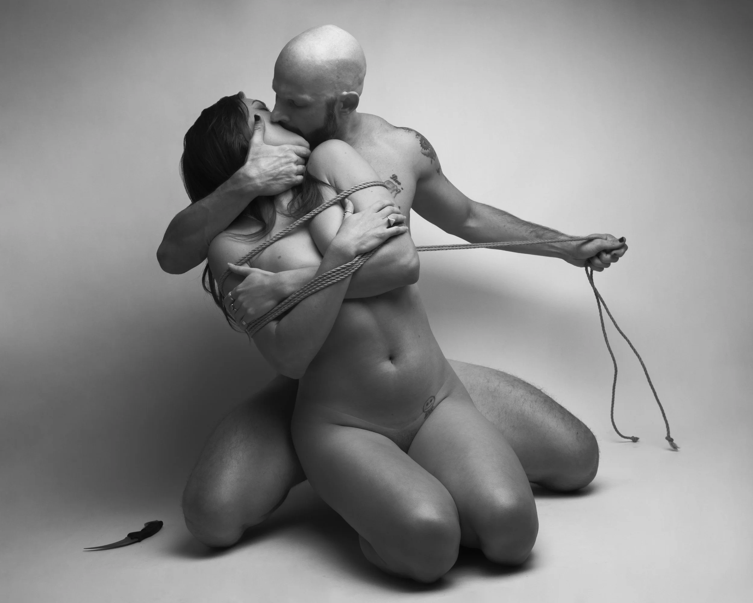 A black and white photo of a man and woman in an intimate pose, with the man holding a rope and the woman being wrapped in it. They are both partially nude, kneeling on the floor, with a plain background.