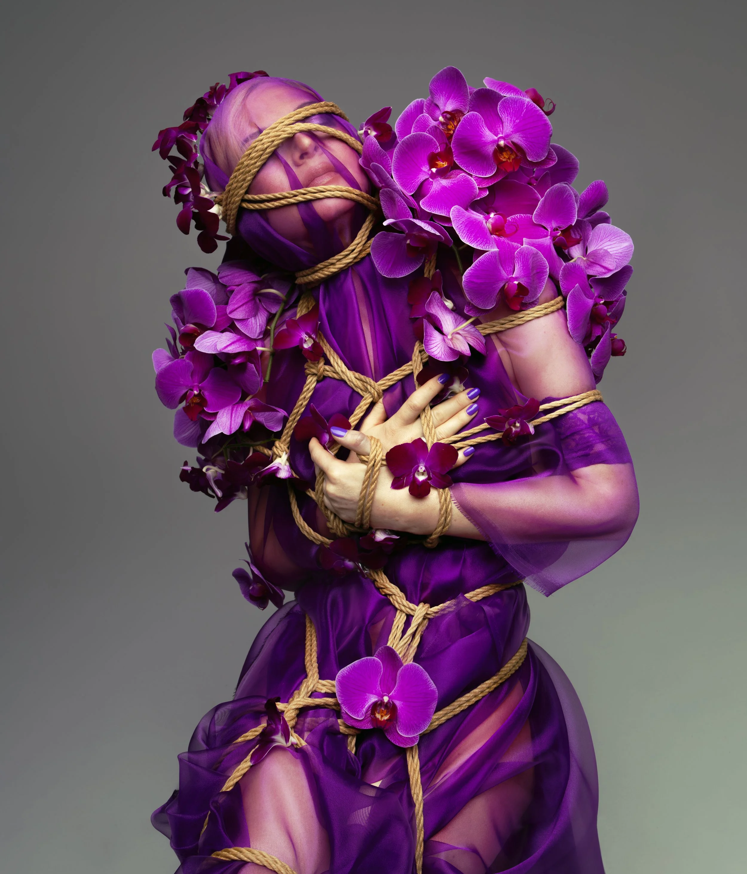 A person wrapped in purple fabric and orchids, with their face mostly obscured by purple ribbons and ropes, posing against a neutral gray background.