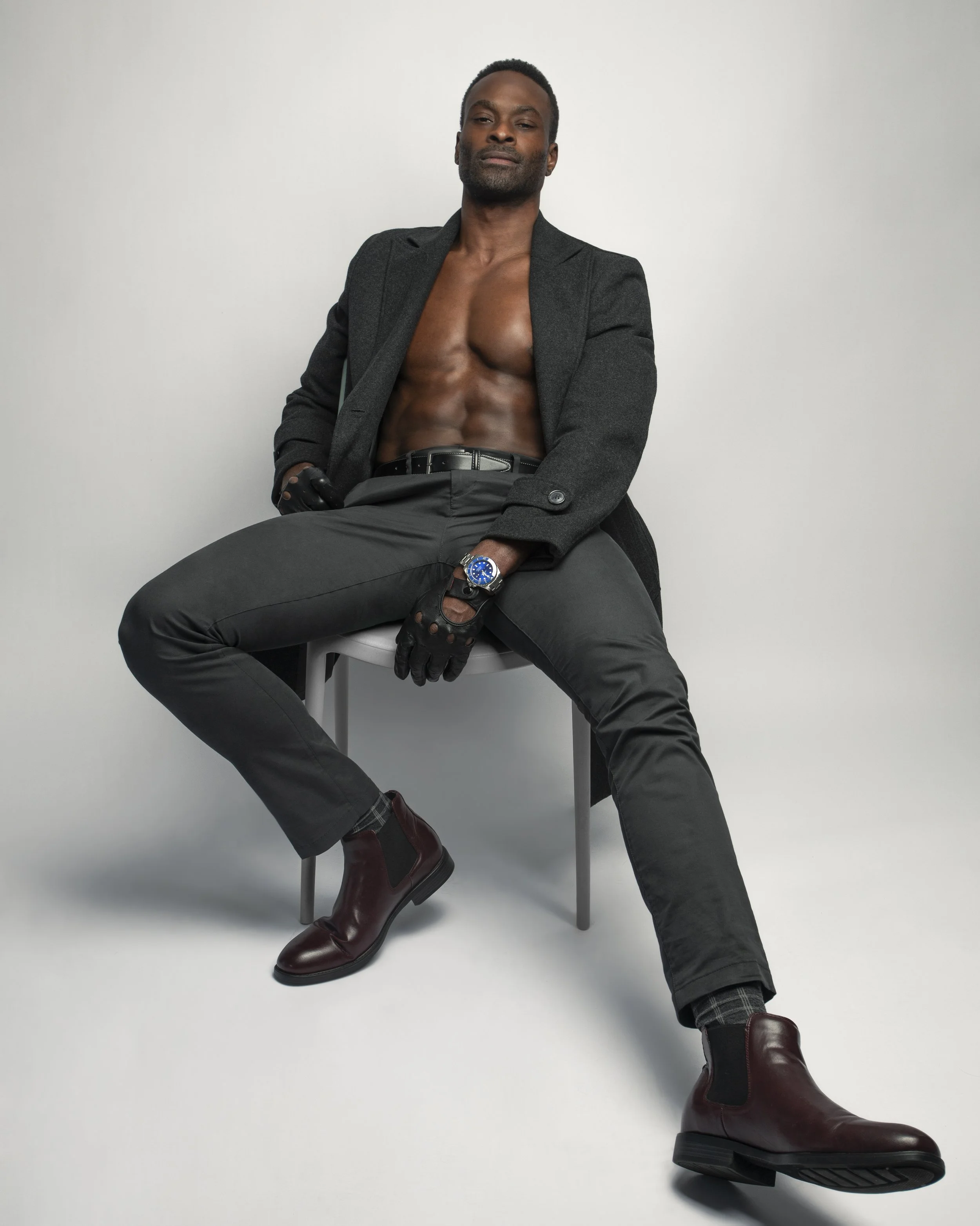 A shirtless man wearing a black blazer, black pants, burgundy boots, and black gloves, sitting on a chair against a white background, with a confident pose.