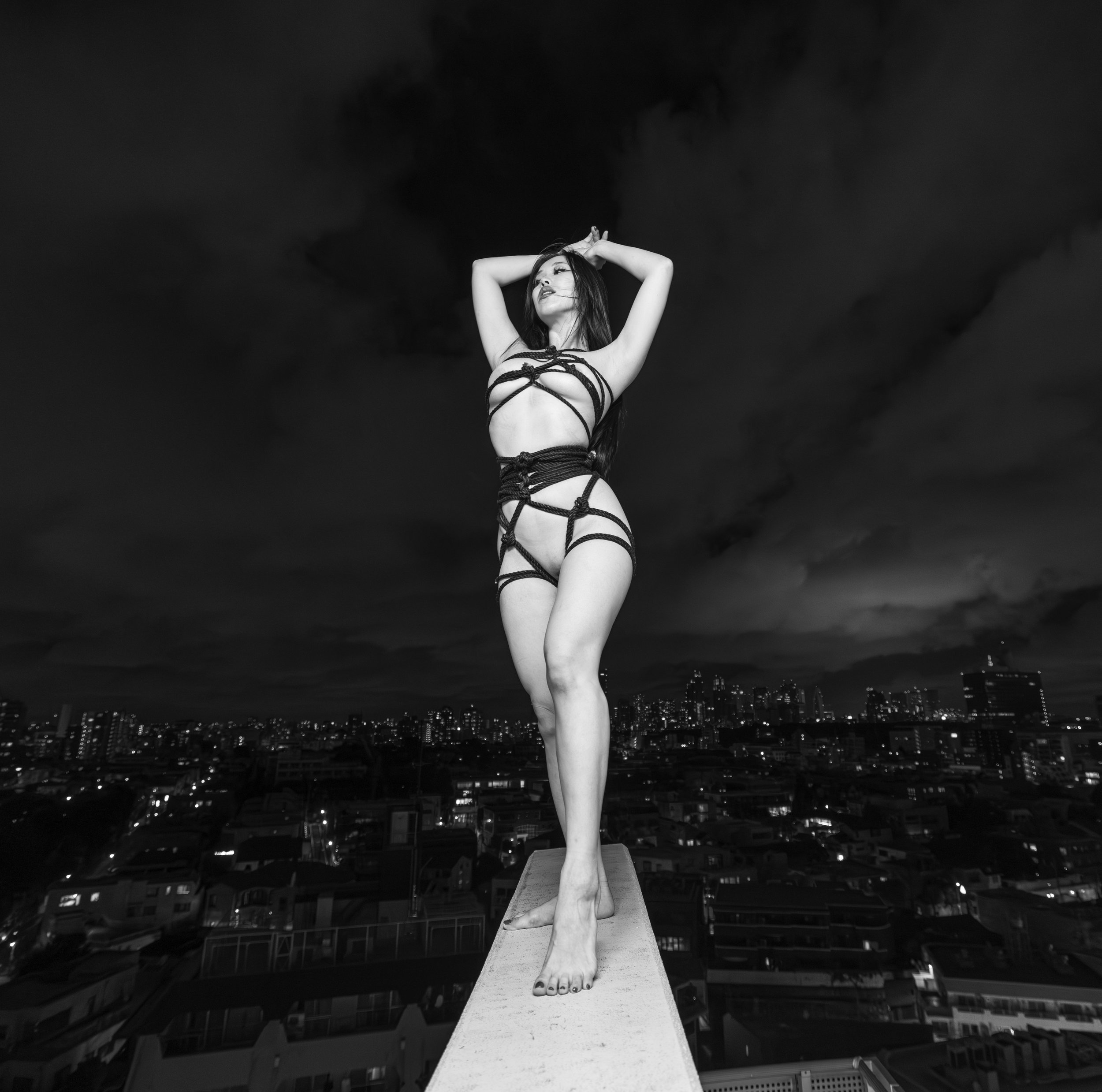 A woman standing on the edge of a rooftop at night, wearing revealing harness-style clothing, with her arms raised above her head and her eyes closed.