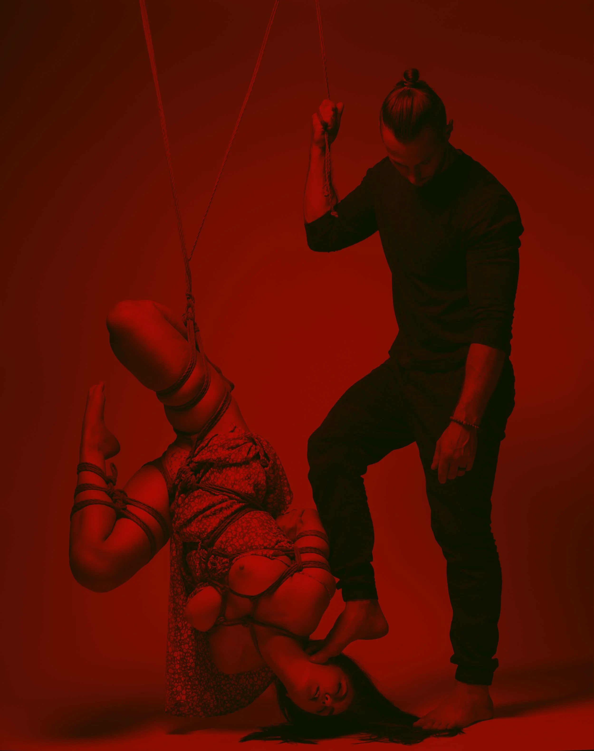A woman is bound and hanging upside down with ropes, while a man stands nearby touching her foot, against a red background.