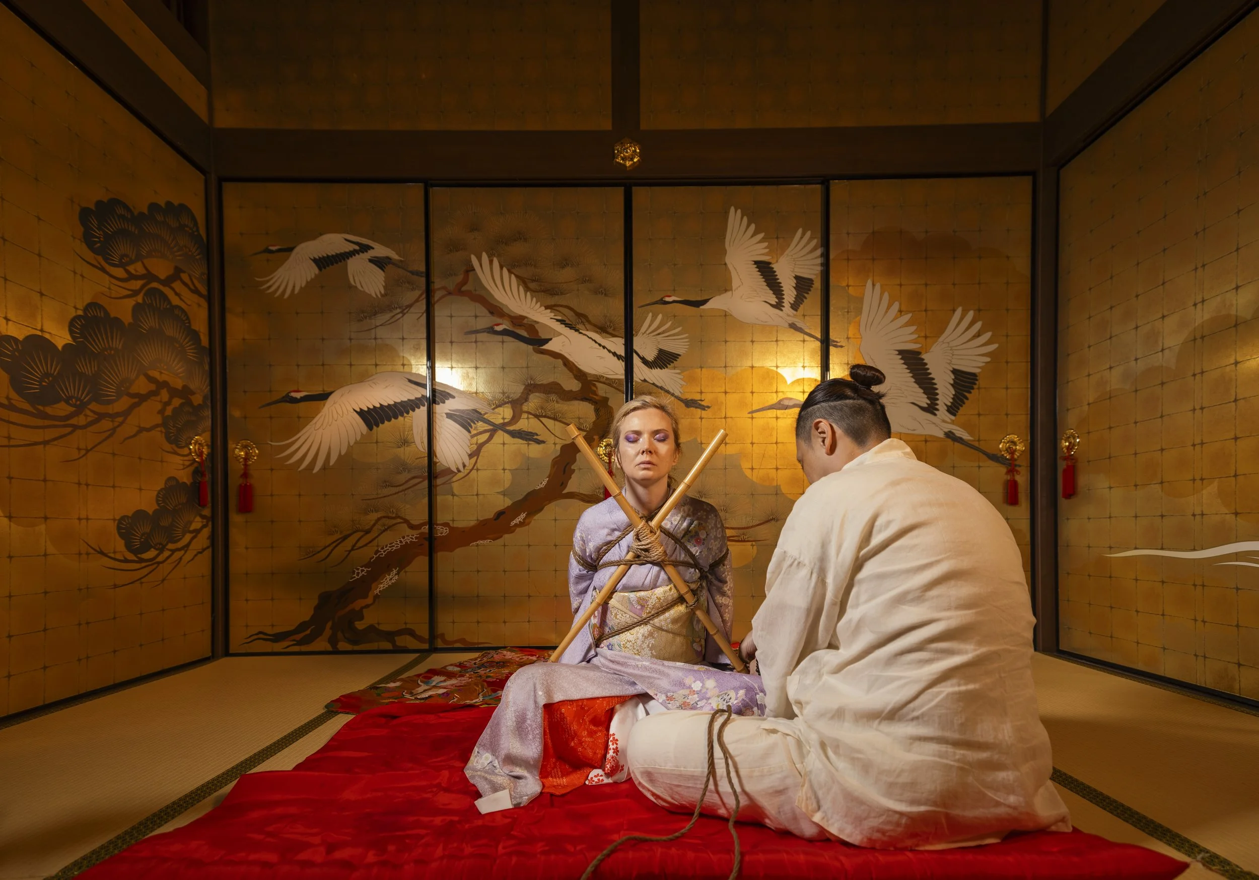 A woman in traditional Japanese attire sits on a red cushion with her hands tied in front of an official, who is also in traditional clothing, in a room with golden folding screens decorated with cranes and trees.