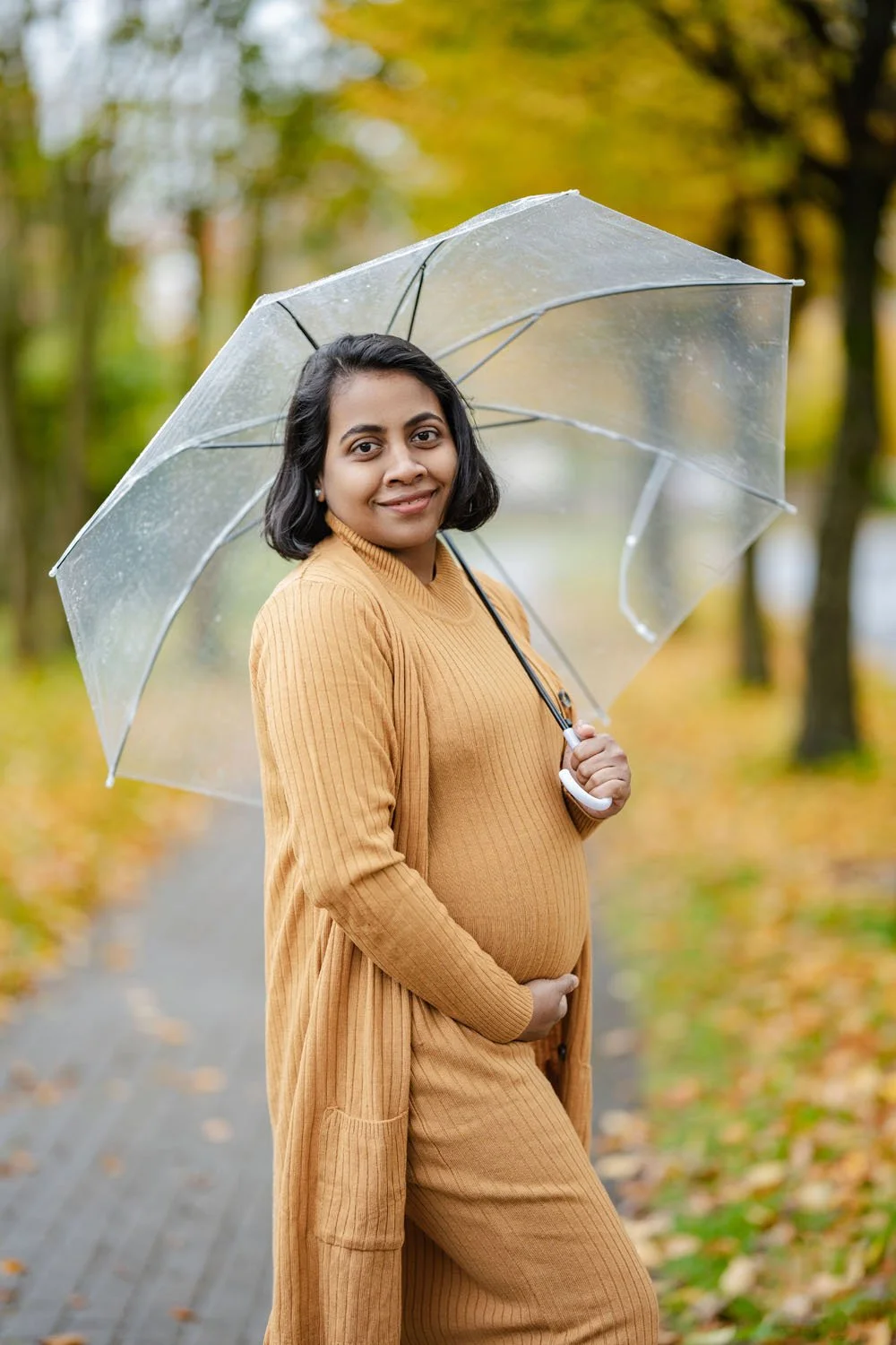 Woman cradling bump in park on rainy day, holding umbrella