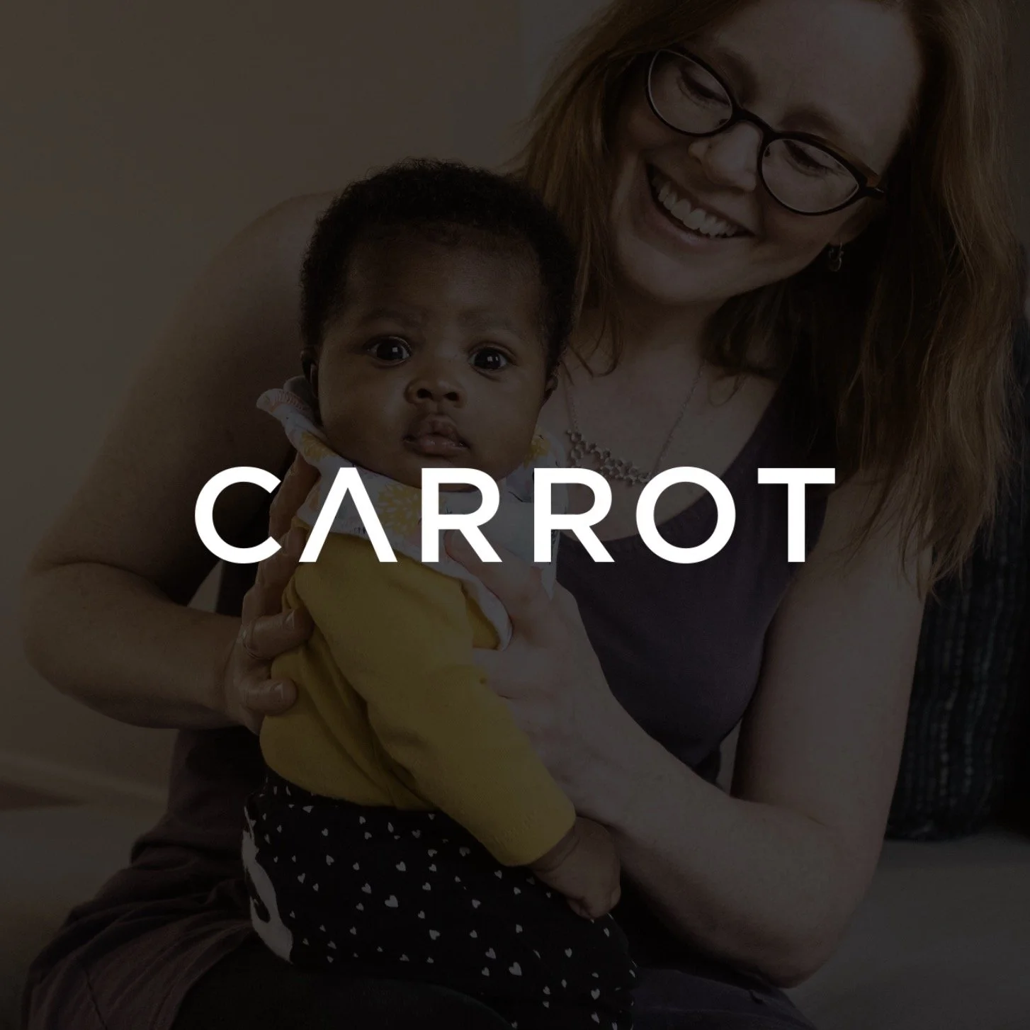 A woman with glasses holding a baby, with a happy expression, in a room with the word 'CARROT' written across the image.