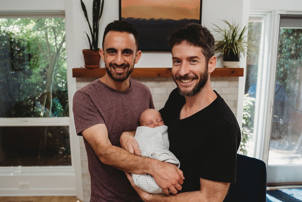 Two men smiling and holding a sleeping baby wrapped in a blanket indoors with a window, plants, and artwork in the background.