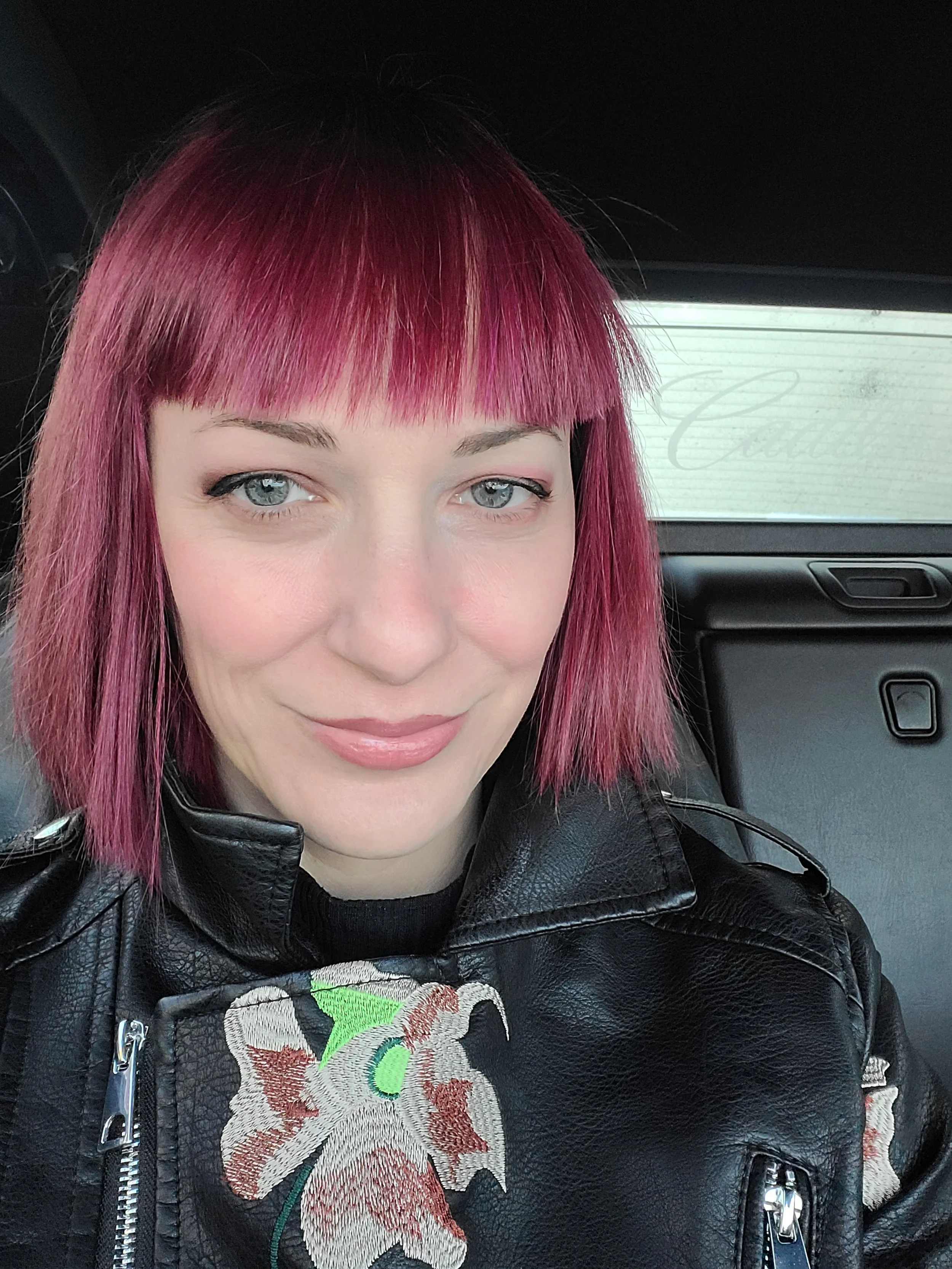 A woman with pink hair and blue eyes, wearing a black leather jacket with floral embroidery, sitting inside a vehicle.