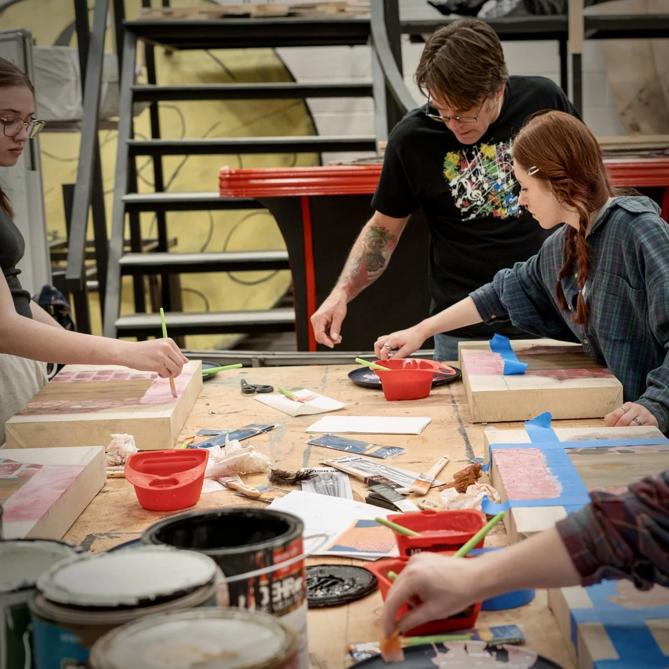 Group of people working on art projects at a cluttered workshop table with paint cans, brushes, and canvases.