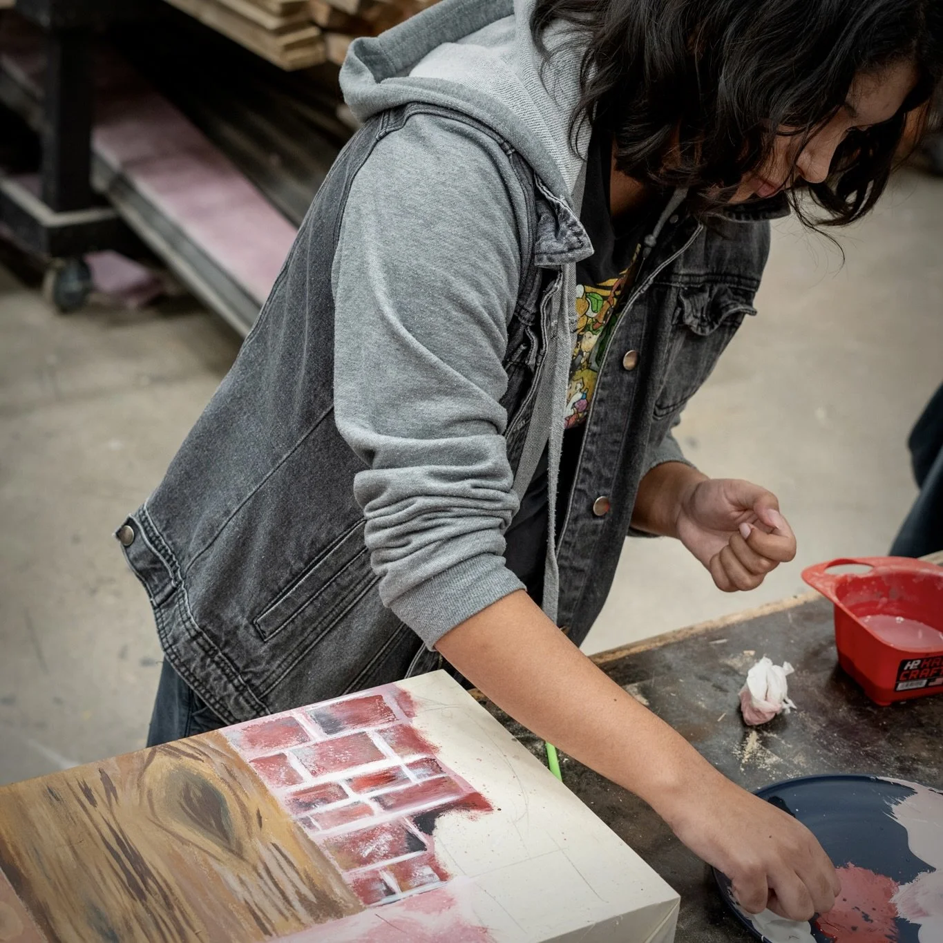 A person with dark hair wearing a gray and black denim jacket working on a painting of a brick wall on a canvas in an art studio.