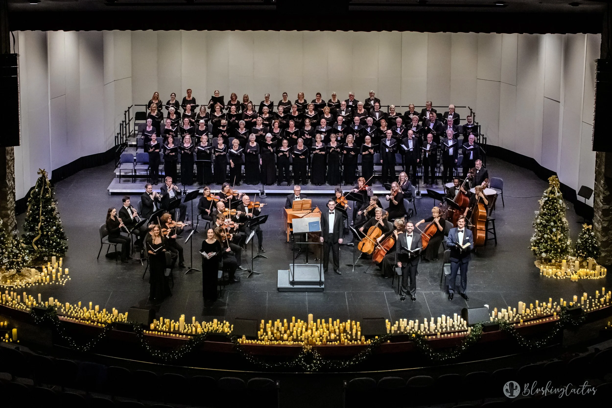 A choir performing on stage with string and wind instrument players, decorated with Christmas trees and candles.