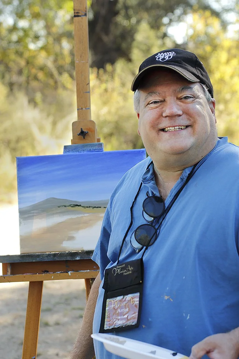 Smiling man wearing a black cap and blue shirt standing outdoors with a painting of a landscape on an easel behind him.