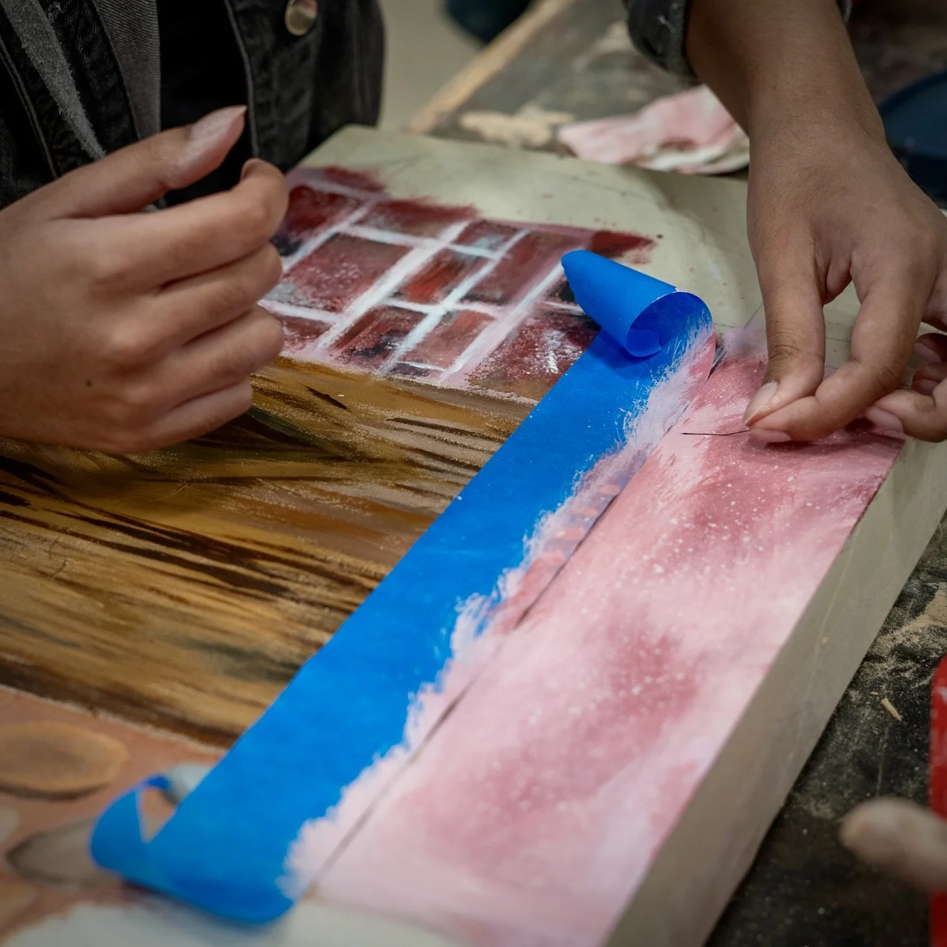 Hands unroll blue painter's tape over a wooden surface with some red and black marks, preparing to peel the tape to reveal a pattern underneath.