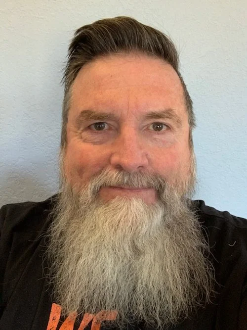Close-up photo of a middle-aged man with brown hair, a full gray beard, wearing a black shirt, standing against a plain light-colored wall.