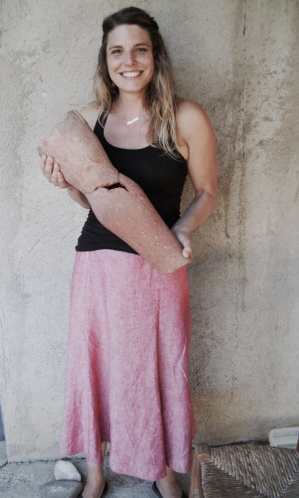 A woman with long curly hair, wearing a black tank top and pink wide-legged pants, smiling and holding a large piece of salt block, standing against a textured concrete wall.