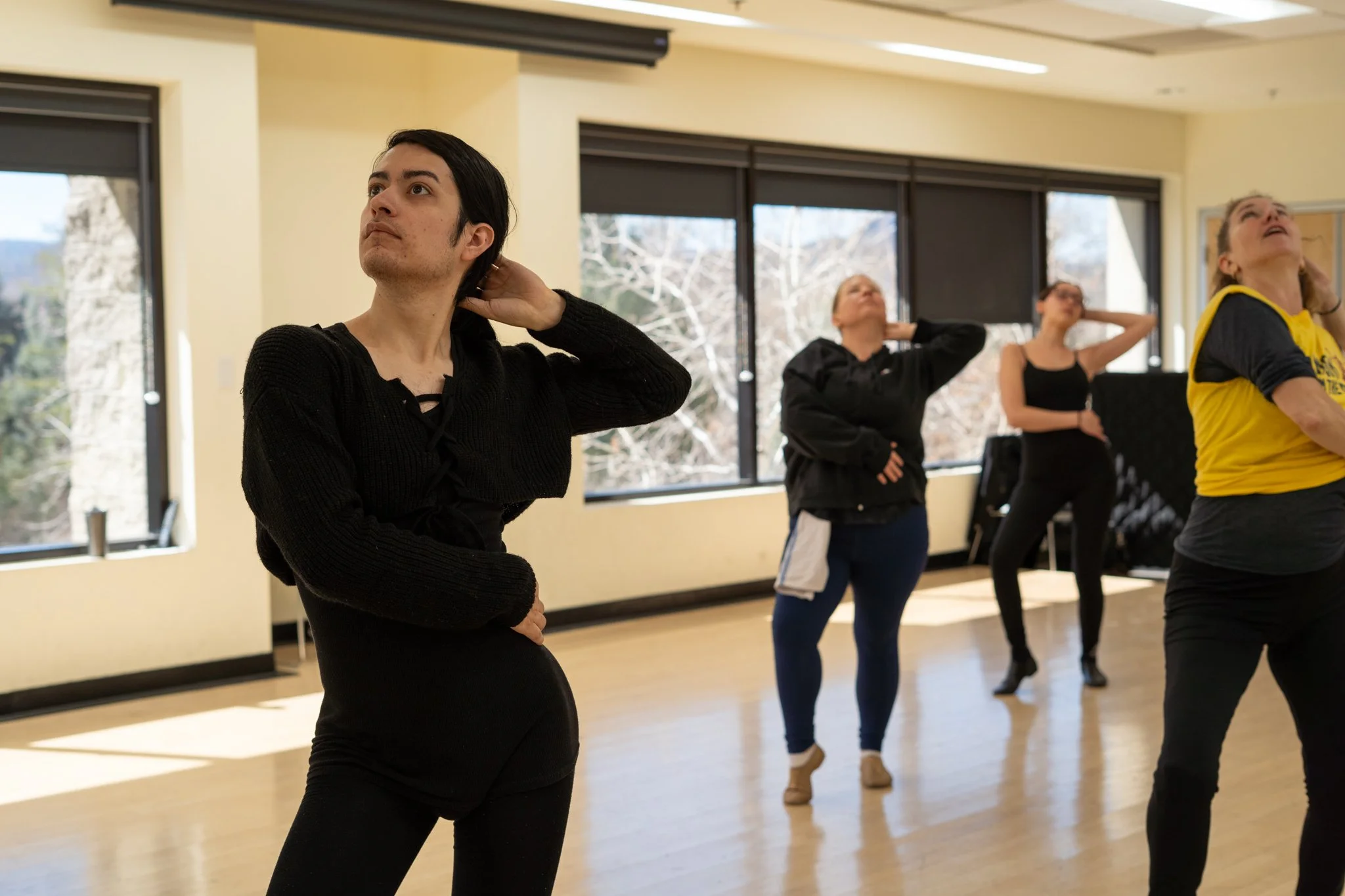 A group of people in a dance studio practicing dance moves, some with their hands behind their heads.