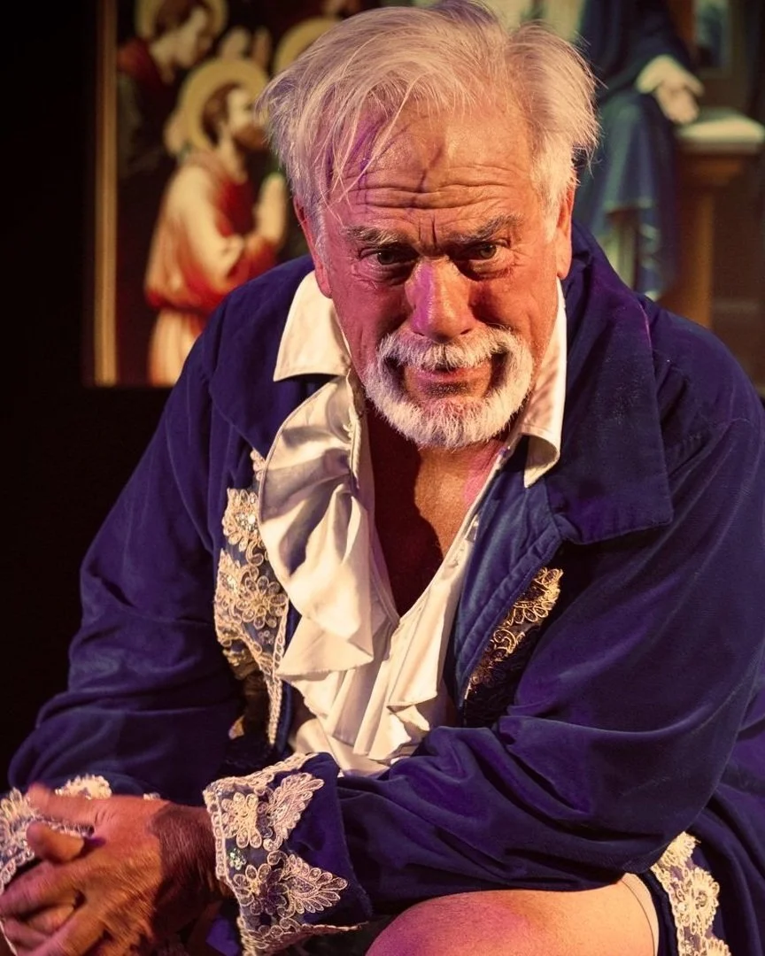 Close-up of an elderly man with white hair and beard, wearing a navy blue jacket with ornate embroidery, sitting with arms crossed, indoor setting with a religious or historical painting in the background.