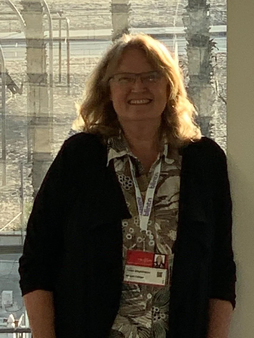 A woman with red hair and glasses smiling, standing indoors near a window during daylight.