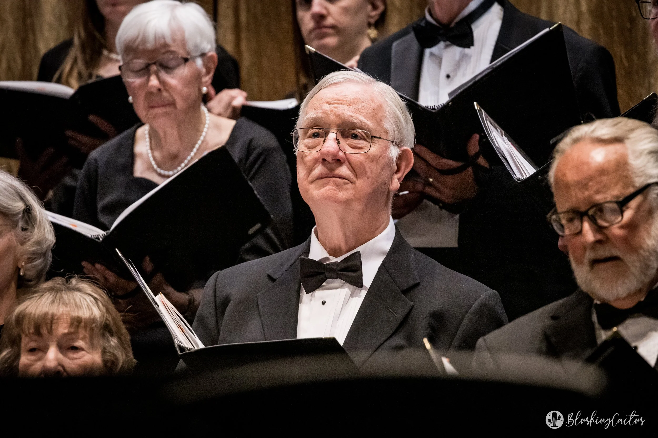 Orchestra members in formal attire, including tuxedos and evening dresses, performing with sheet music in front of them during a concert.