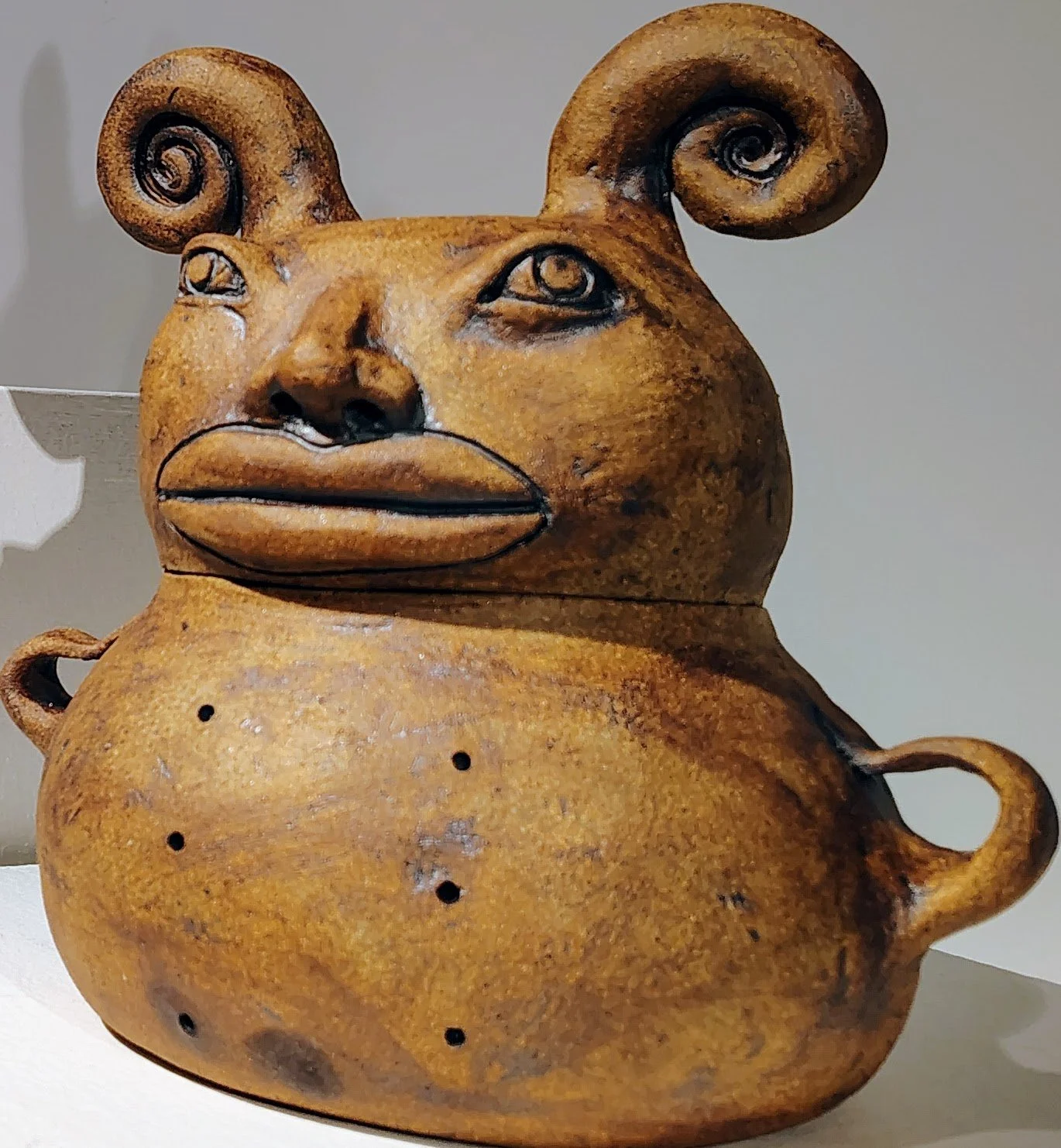 A ceramic sculpture of a creature with a ram's head and human body, featuring curled horns, human-like eyes, a nose, and pursed lips, with a rounded body and small holes.