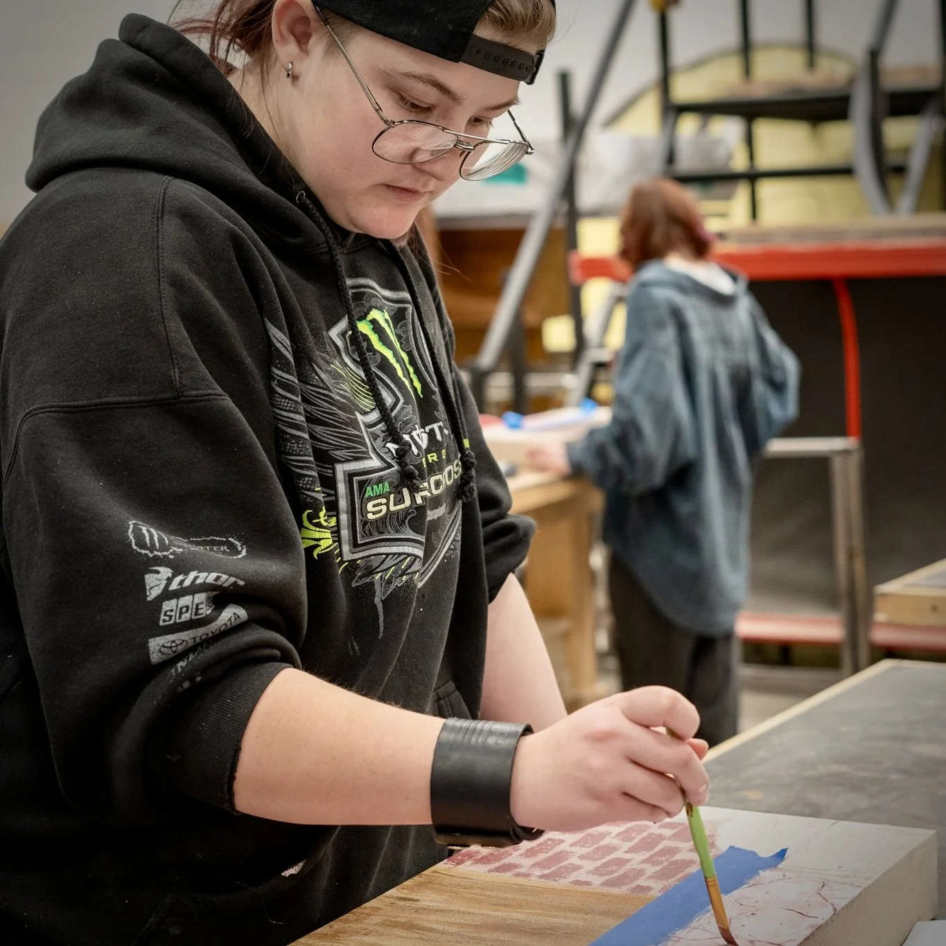A person with glasses and a backwards cap painting on a piece of paper or canvas on a wooden table, with a person in the background in a workshop or classroom setting.