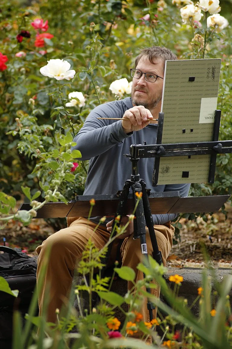 A man with glasses, a beard, and a long-sleeved gray shirt is painting outdoors among blooming roses with white and pink flowers. He is sitting on a rock, holding a paintbrush in his right hand, and facing an easel with a palette or sketchpad. There