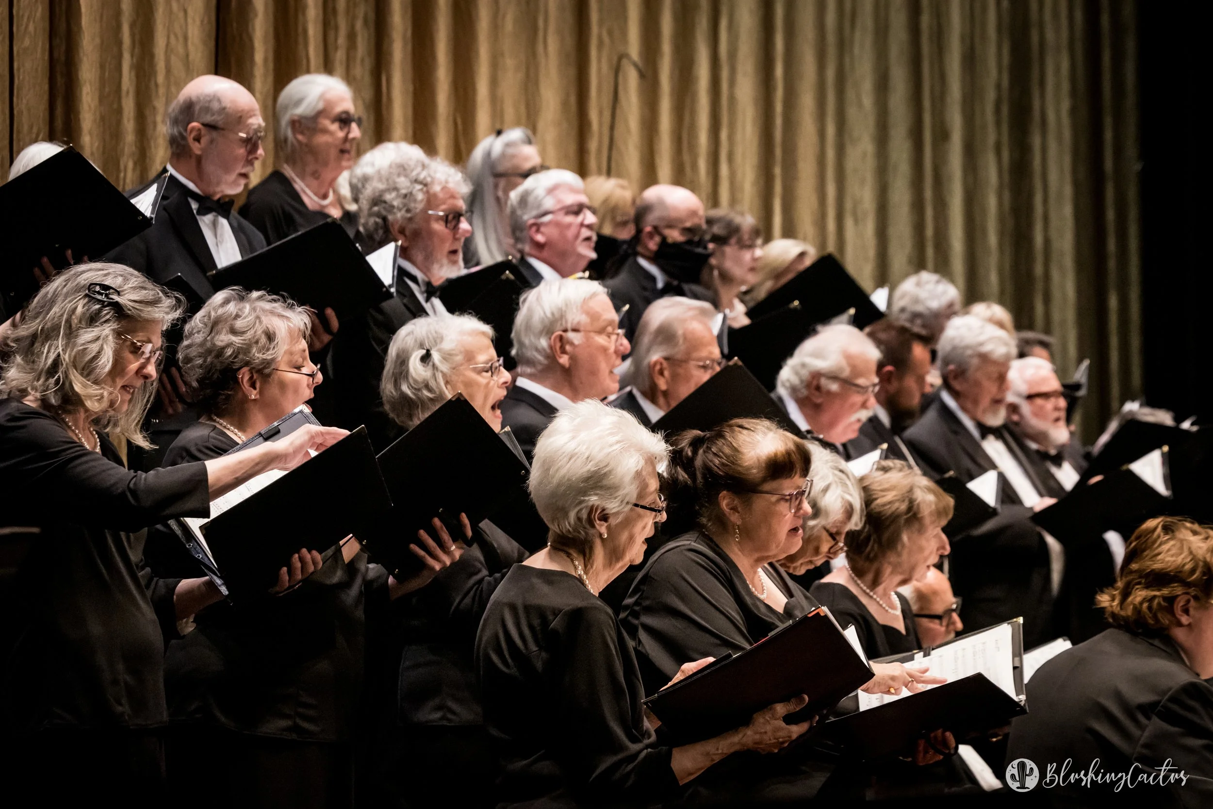 Choir of elderly men and women singing with black folders, dressed in black formal attire, performing on stage with gold curtains in the background.