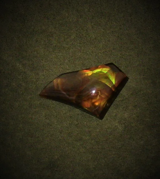 A polished, irregularly-shaped stone with iridescent colors lies on a dark, textured surface.