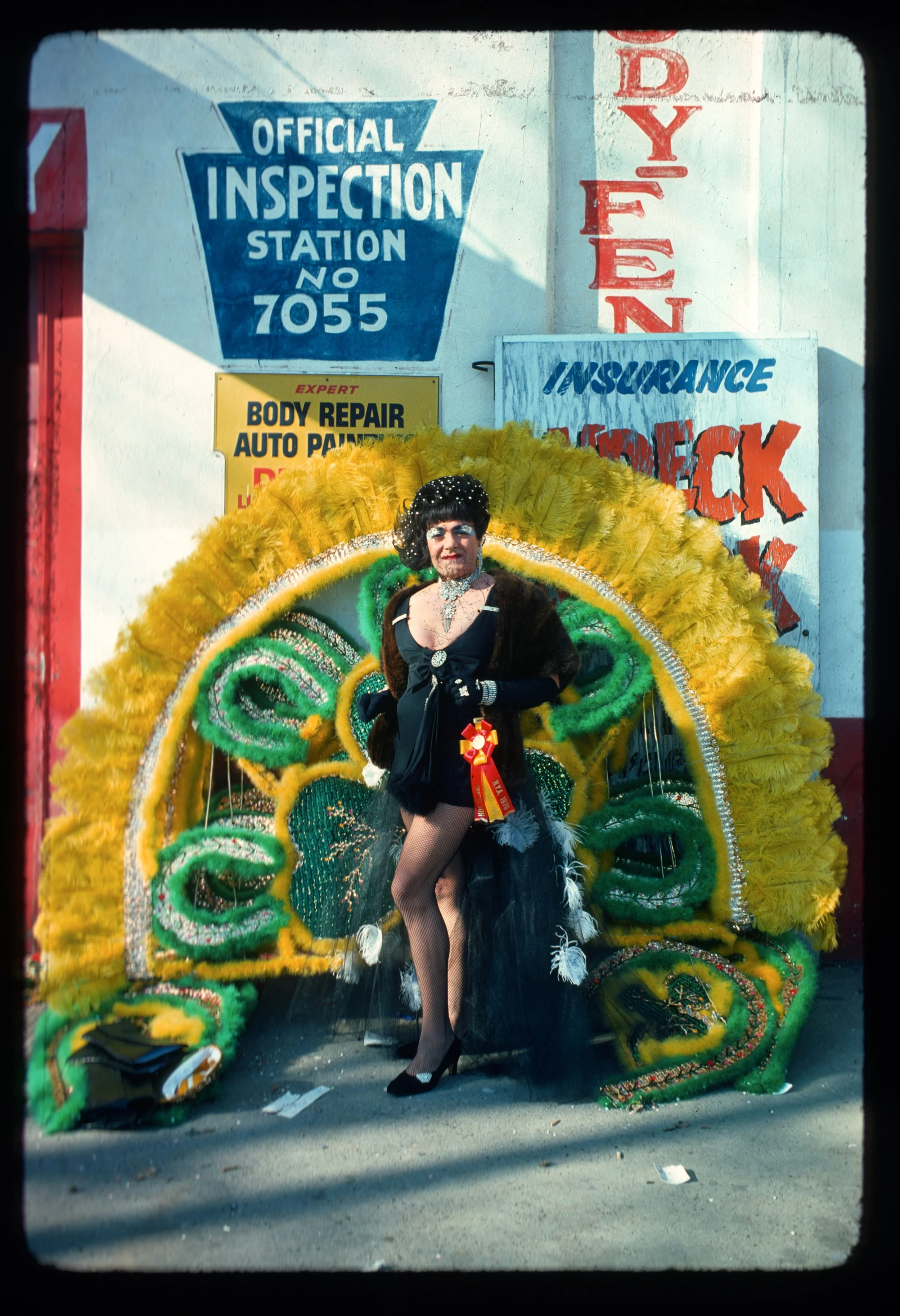 Person dressed in theatrical costume standing in front of a colorful parade float, with signs for an auto body repair shop, insurance, and a DYLAN sign in the background.