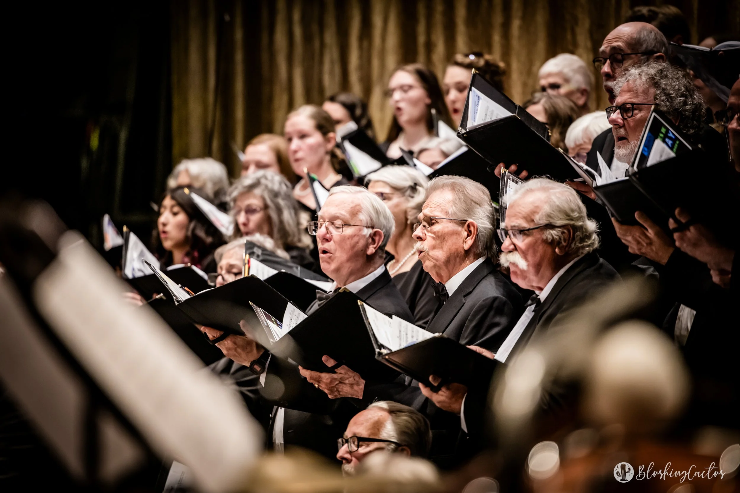 A choir performing on stage, dressed in formal black attire, holding sheet music, with a golden curtain background.