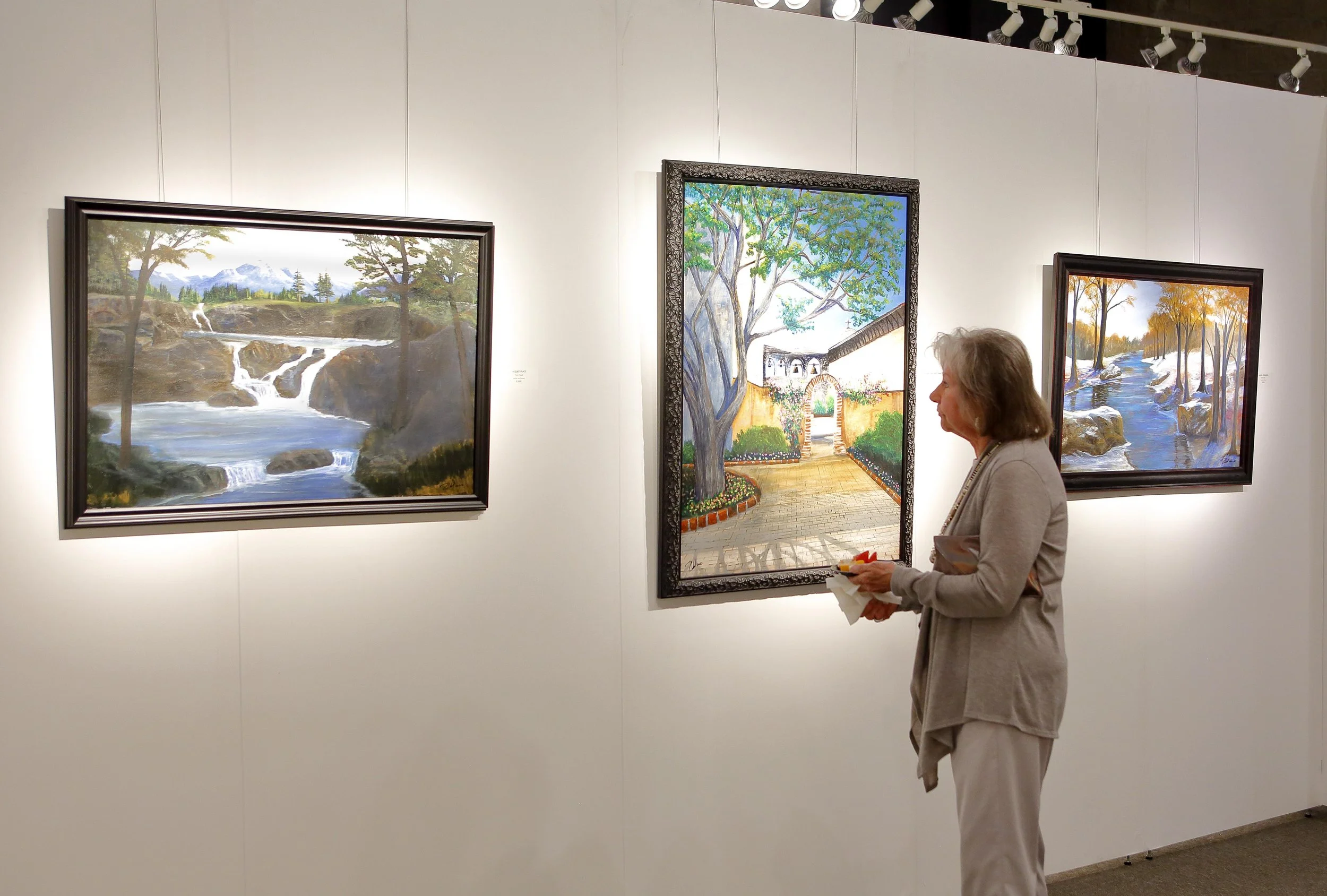 An elderly woman viewing paintings in an art gallery. Three landscape paintings are visible on the wall, featuring natural scenes such as waterfalls, trees, and a river.